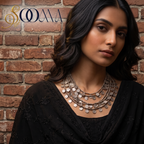 A woman with dark hair models a multi-layered silver coin necklace against a rustic red brick wall background. She is wearing a black embroidered top and a black dupatta.