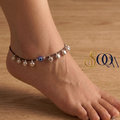 Side view of foot wearing black thread evil eye anklet with silver ghungroo drops on dark wooden floor