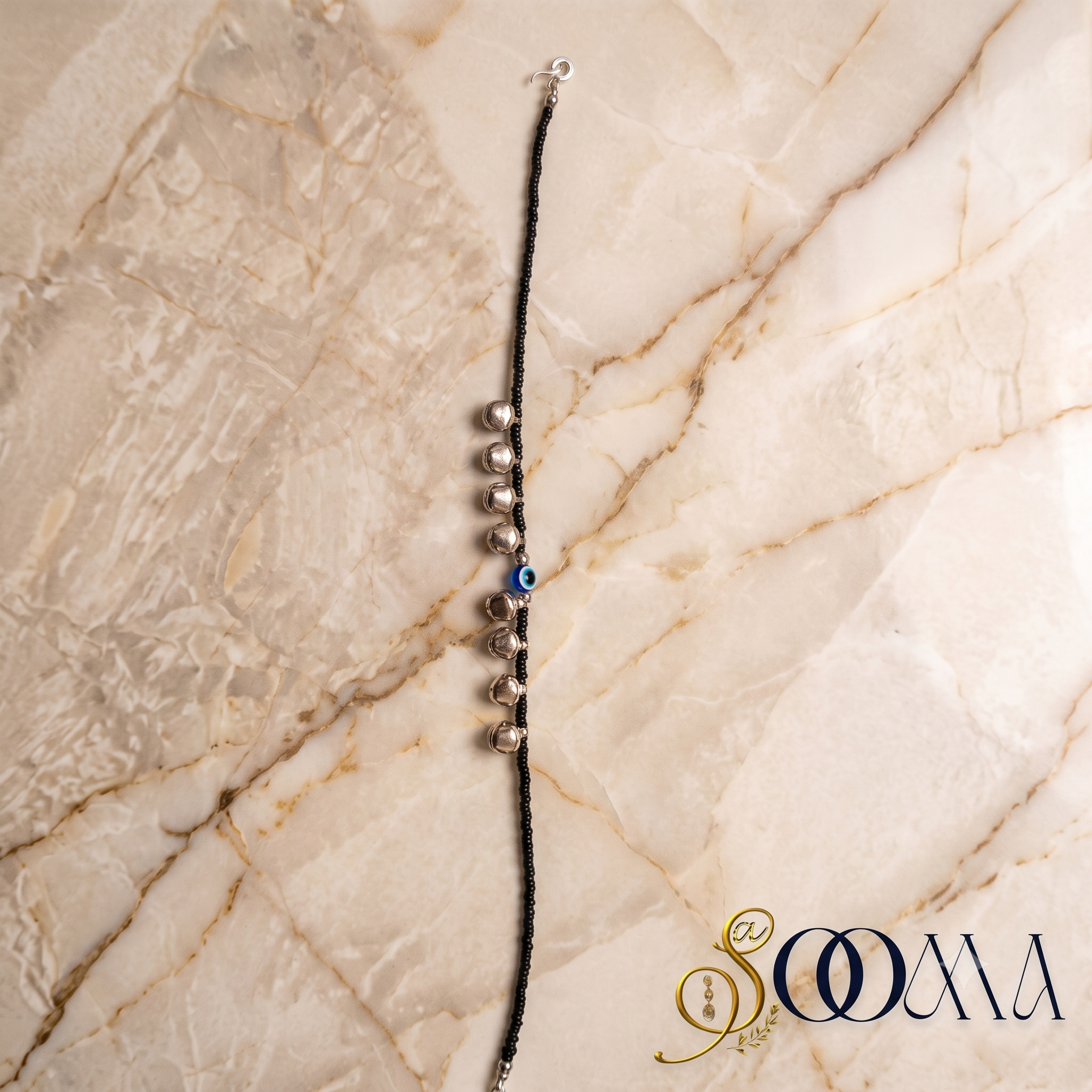 Sooma brand black bead evil eye anklet with silver ghungroo bells displayed flat on marble