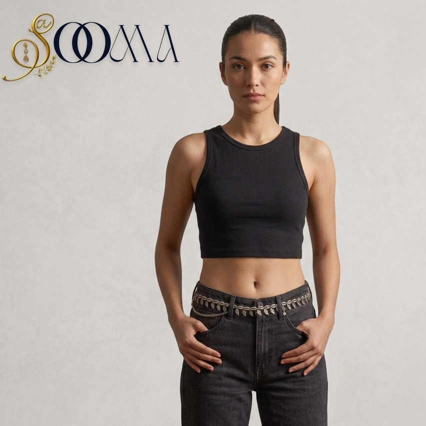 A woman stands against a light textured wall wearing a sleeveless black crop top and dark jeans with a decorative metallic chain detail at the waist. The brand name 'SOMA' appears in the top left corner.