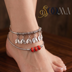 A pair of silver-toned anklets worn on a person's ankle and foot, featuring dangling leaf charms and a red beaded accent, shown against a dark wooden background.