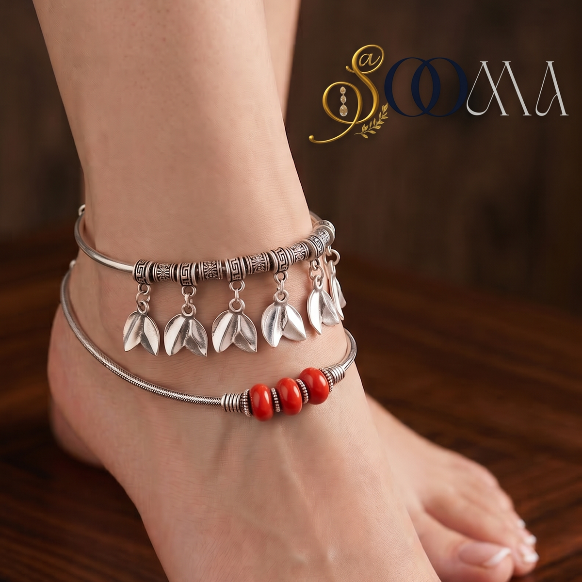 A pair of silver-toned anklets worn on a person's ankle and foot, featuring dangling leaf charms and a red beaded accent, shown against a dark wooden background.
