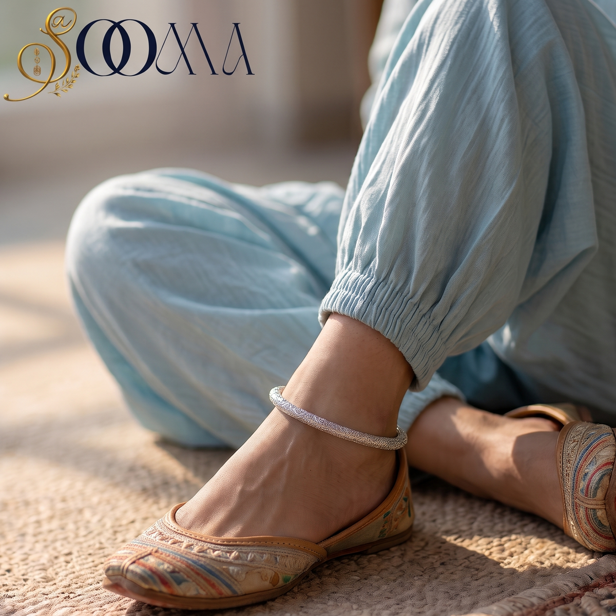 A person wearing light blue loose pants and complimenting ankle kada  sits on a woven rug. The brand name 'SOOMA' is visible in the upper left corner.