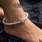 A sterling silver bangle anklet with intricate floral engravings and a central decorative clasp, shown worn on a person's ankle. The brand logo 'SOOMA' is visible in the upper left, and the background is a dark plush fabric.