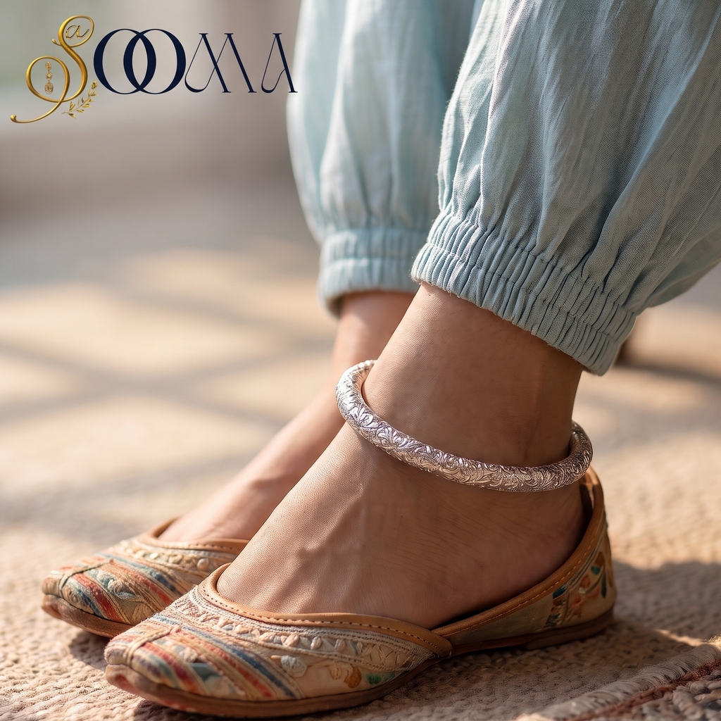 A close-up lifestyle shot of a person wearing light blue gathered pants and embroidered flat shoes with a silver anklet, standing on a textured woven rug. The brand logo 'SOOMA' is visible in the top left corner.