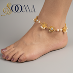 A gold-toned floral anklet with small white pearl-like beads worn on a person's ankle, shown resting on a wooden surface with a neutral fabric background. The brand name SOOMA appears in the upper left.
