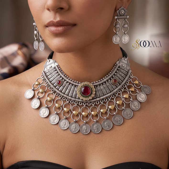 A close-up of a model wearing an ornate silver-toned statement necklace with dangling coin charms, red gemstone accents, and gold-toned heart-shaped details, paired with matching drop earrings.