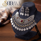 An ornate silver-toned tribal-style necklace and matching drop earrings displayed on a dark velvet jewelry bust. The necklace features multiple dangling coin-shaped charms, beaded fringe, and red gemstone accents, with a black tassel hanging from the base.