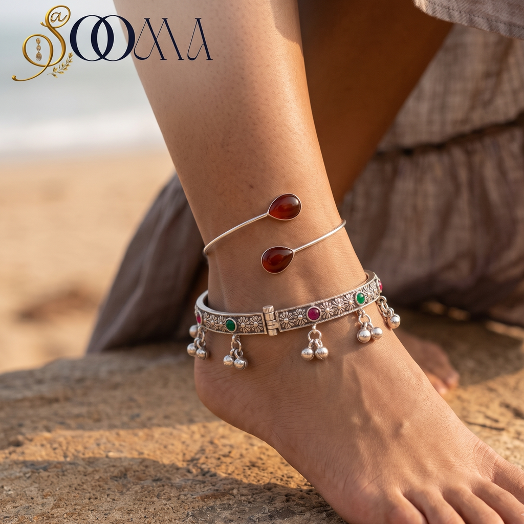 A close-up lifestyle shot of a person's ankle and foot wearing a pair of silver-toned Indian-style anklets on a sandy beach. The anklets feature silver chains with small green and pink stones, red stones, and dangling silver bells.