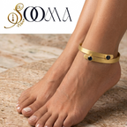 A gold-toned wrap-style ankle bracelet with two small red gemstone accents, shown worn on a person's ankle with bare feet on a beige stone surface. The SOOMA brand logo appears in the top left corner.