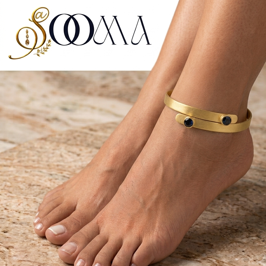 A gold-toned wrap-style ankle bracelet with two small red gemstone accents, shown worn on a person's ankle with bare feet on a beige stone surface. The SOOMA brand logo appears in the top left corner.