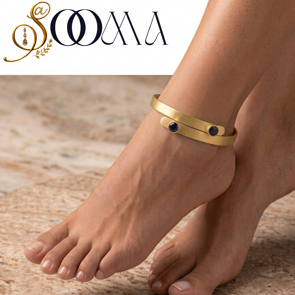 A pair of bare feet wearing a slim gold cuff anklet with two small dark oval stones, standing on a beige stone floor. The brand logo 'SOMA' is visible in the top left corner.