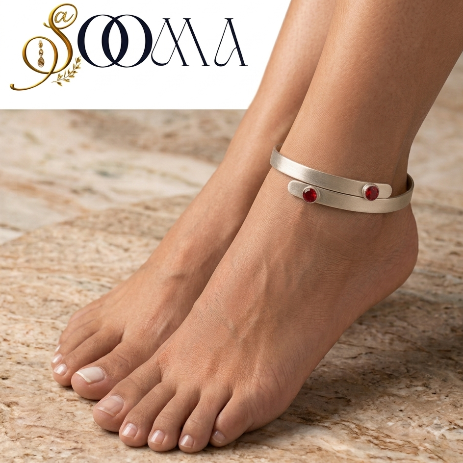 A silver-toned wrap-style ankle bracelet with two small red gemstone accents, shown worn on a person's ankle with bare feet on a beige stone surface. The SOOMA brand logo appears in the top left corner.