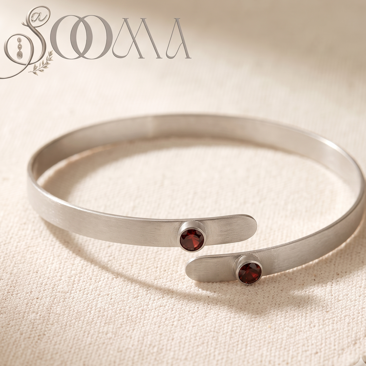 A silver-toned open cuff bracelet with two small round dark red gemstone accents, displayed on a textured beige fabric background. The brand name 'SOAA' is visible in the top left corner.