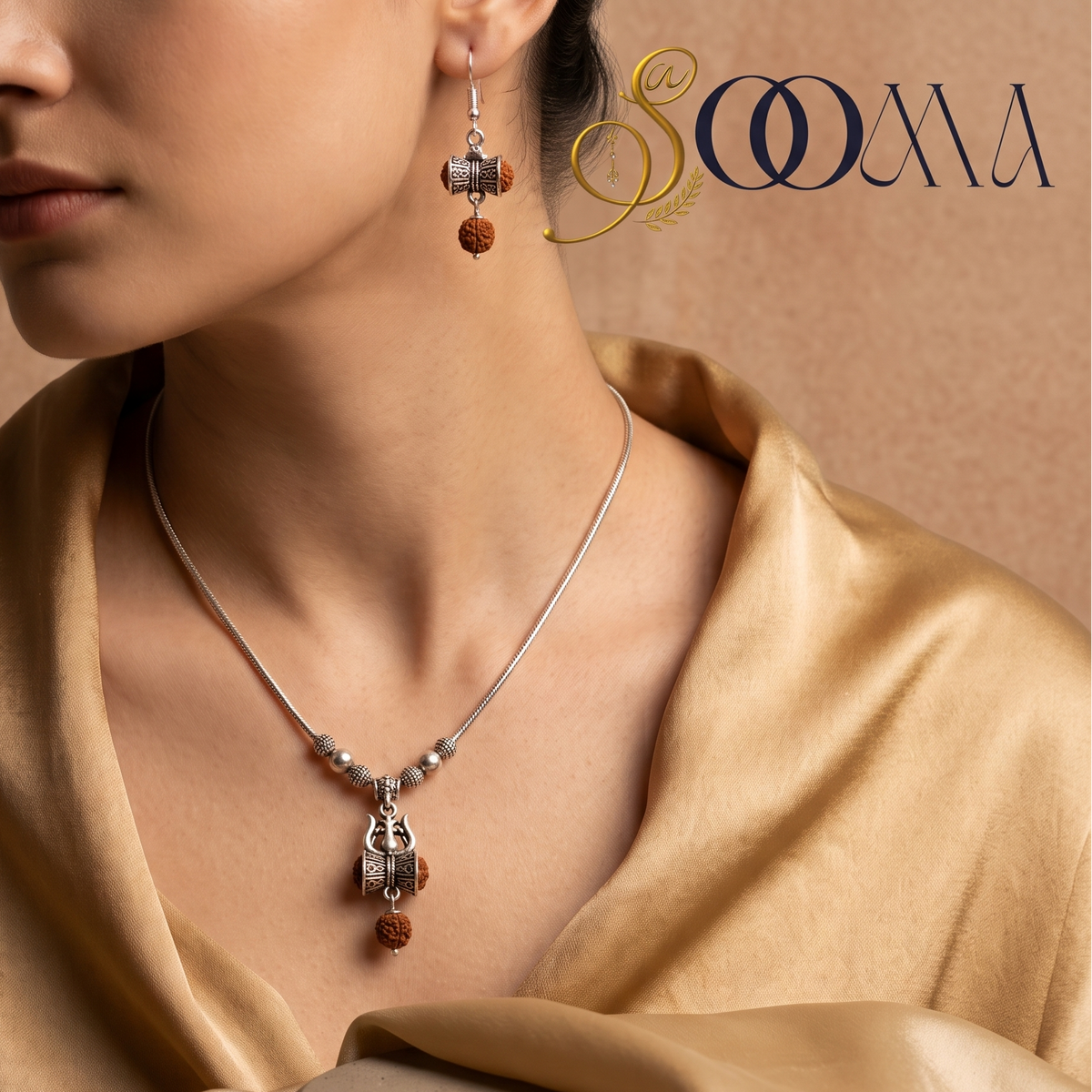 A close-up of a woman wearing a delicate silver necklace with a rudraksha pendant and matching dangling earrings, styled with a gold satin shawl against a warm beige background. The brand logo 'SOOMA' is visible in the top right corner.