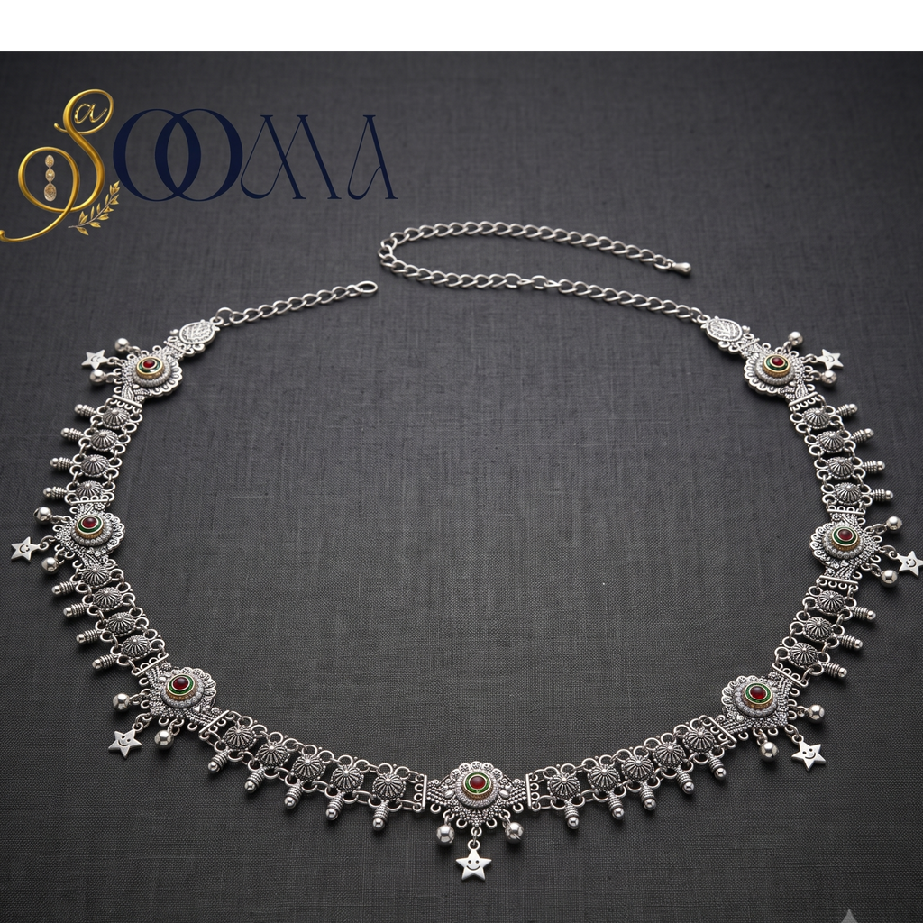 An ornate silver-toned belly chain necklace with intricate filigree, dangling star charms, and small amber-colored stone accents, displayed on a dark textured background with the SOOMA brand logo in the top left corner.