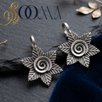 A pair of silver-toned starburst ear cuff with a central spiral design, displayed on a dark slate surface next to a deep blue velvet cloth. The brand logo 'SOMMA' is visible in the top left corner.