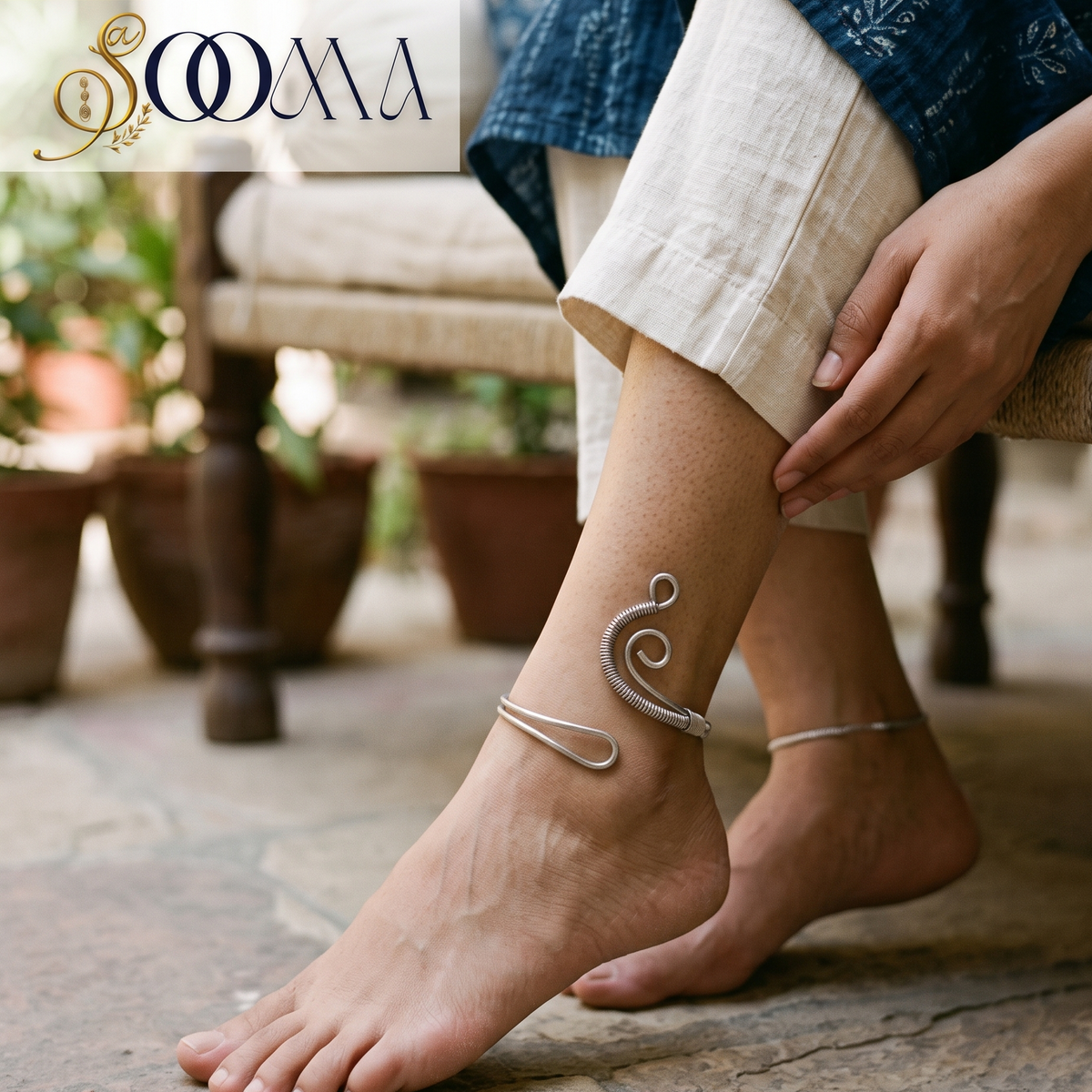 Side perspective view of model seated indoors wearing a wire-wrapped spiral wave silver ankle cuff paired with linen trousers