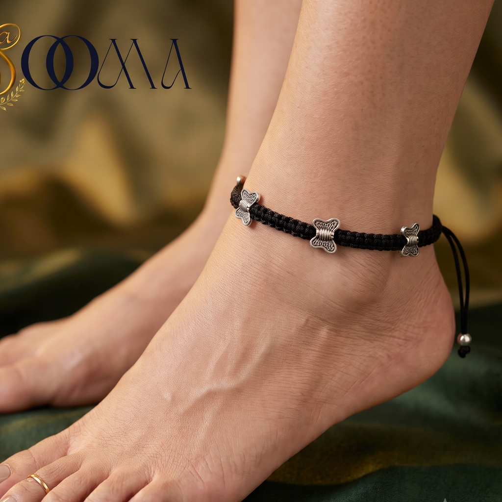 A close-up lifestyle shot of a person's feet wearing a black braided ankle bracelet with silver bone charms, set against a dark green draped fabric background with a gold logo overlay.