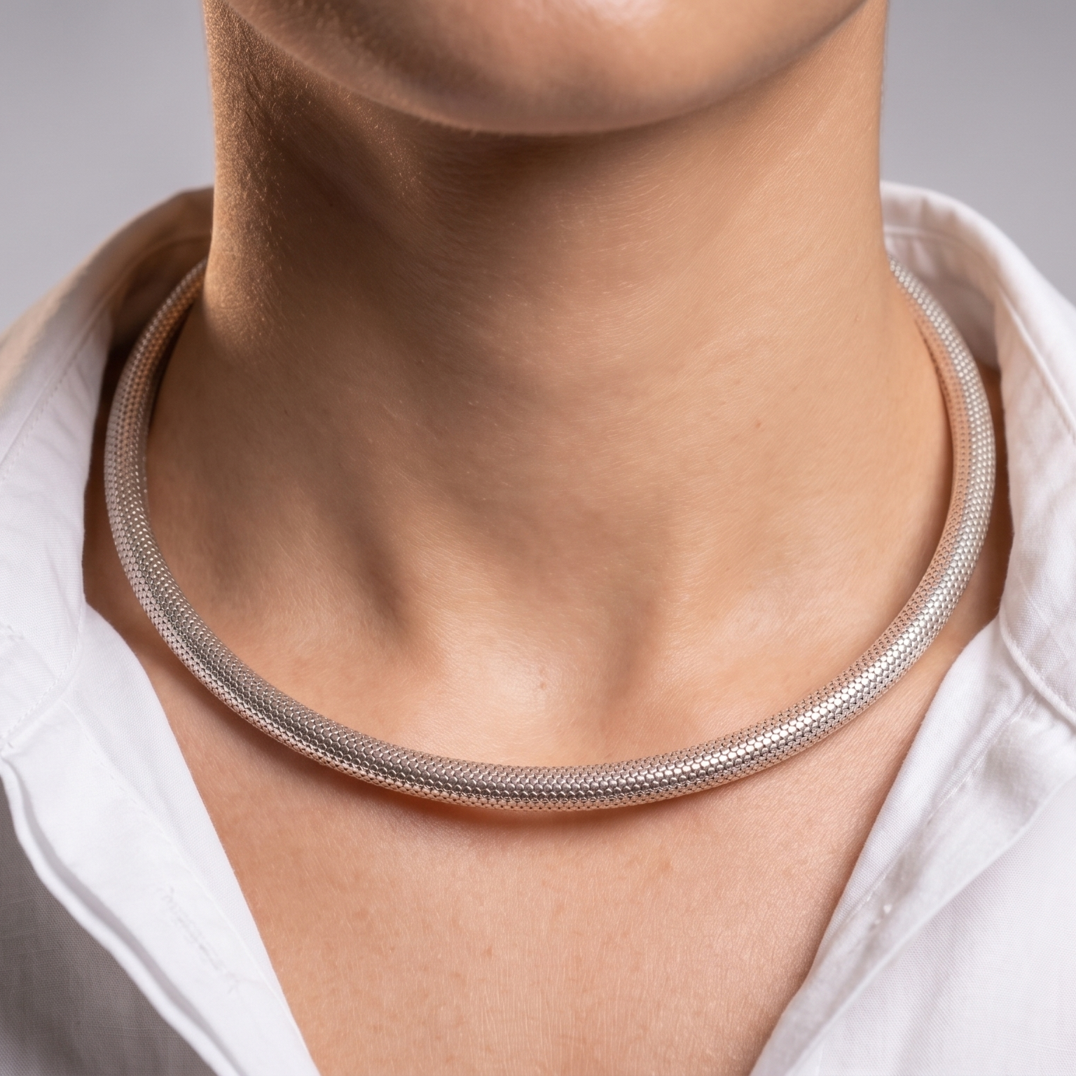 A close-up of a person's neck and collarbone area wearing a slim silver snake chain hasli necklace, paired with a white collared shirt against a soft gray studio background.