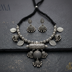 An antique silver-toned jewelry set featuring a necklace with a large cylindrical pendant, leaf-shaped beads, and matching jhumka earrings, displayed on a dark textured slate background. The brand logo 'SOOMA' is visible in the top left corner.