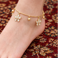 A close-up of a person's foot and ankle wearing a delicate gold anklet with flower charms and small bead drops, standing on a rocky outdoor surface. The brand name 'SOOMA' is visible in the top left corner.
