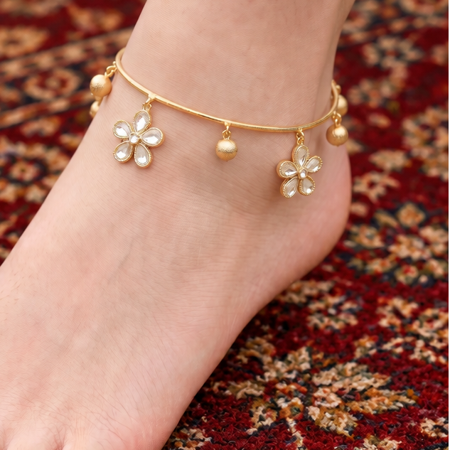 A close-up of a person's foot and ankle wearing a delicate gold anklet with flower charms and small bead drops, standing on a rocky outdoor surface. The brand name 'SOOMA' is visible in the top left corner.