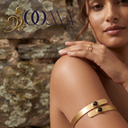 A woman wearing a gold cuff bracelet with two small black oval stones, posed against a rustic stone wall. The brand logo 'SOMA' is visible in the top left corner.