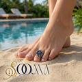 Model wearing an adjustable oxidized silver floral toe ring with deep blue stones on a sandy beach next to a swimming pool