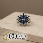 Close up of Sooma brand intricate eight petal silver flower toe ring featuring midnight blue stones on a beige concrete texture