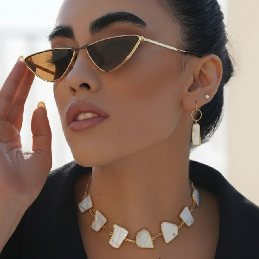 Woman wearing cat-eye sunglasses, gold and white stone necklace and earrings with a neutral background
