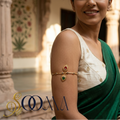 A woman wearing a gold-toned armlet bangle with red and green stones on her upper arm, dressed in a white embroidered blouse and green sari, standing in an ornate indoor courtyard. The brand name 'SOOMA' is displayed in the lower-left corner.