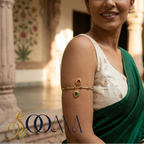 A woman wearing a gold-toned armlet bangle with red and green stones on her upper arm, dressed in a white embroidered blouse and green sari, standing in an ornate indoor courtyard. The brand name 'SOOMA' is displayed in the lower-left corner.