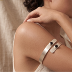 A woman wearing two silver cuff bracelets with small black oval stones, posed against a soft beige fabric backdrop. The brand logo 'soama' is visible in the top left corner.