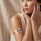 A close-up lifestyle shot of a model wearing minimalist silver cuff bracelets with black oval centers on her upper arm, against a soft beige linen background.