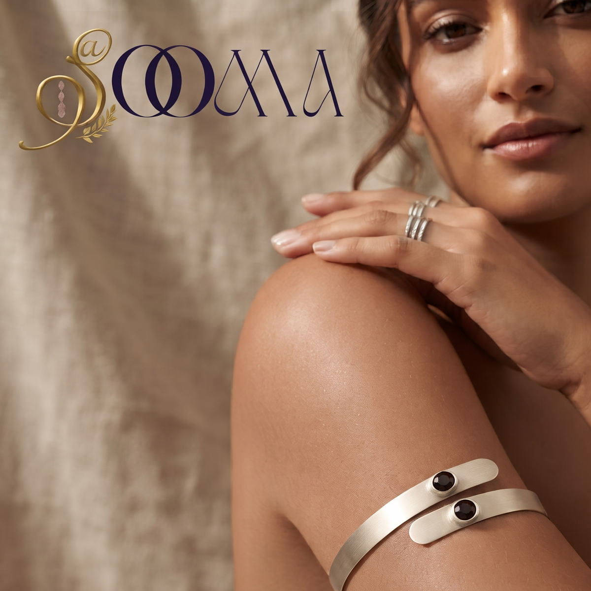 A woman wearing a wide silver cuff bracelet with two small black oval stones on her upper arm, with matching silver rings on her fingers. The brand logo 'SOOMA' is visible in the top left corner against a textured beige backdrop.
